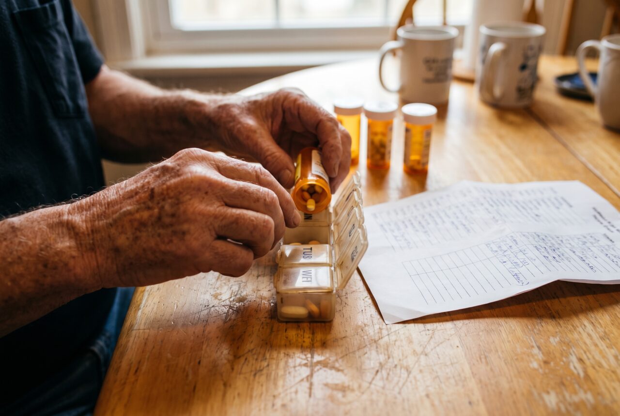 Older hands sorting prescription bottles into a weekly pill organizer to build a two-week hurricane medication supply