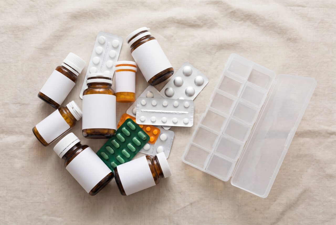 Overhead view of many prescription bottles beside an empty weekly pill organizer illustrating polypharmacy in seniors