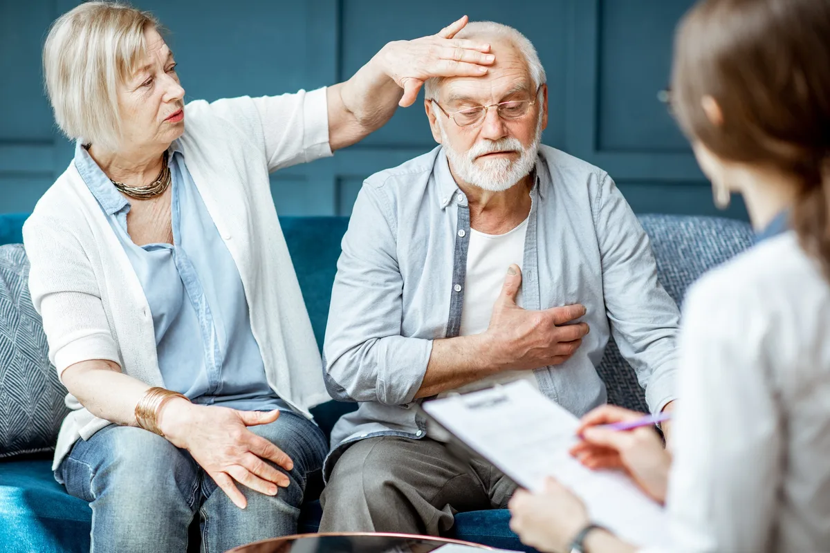 An older couple sit together on a sofa and talk with a clinician during a consultation