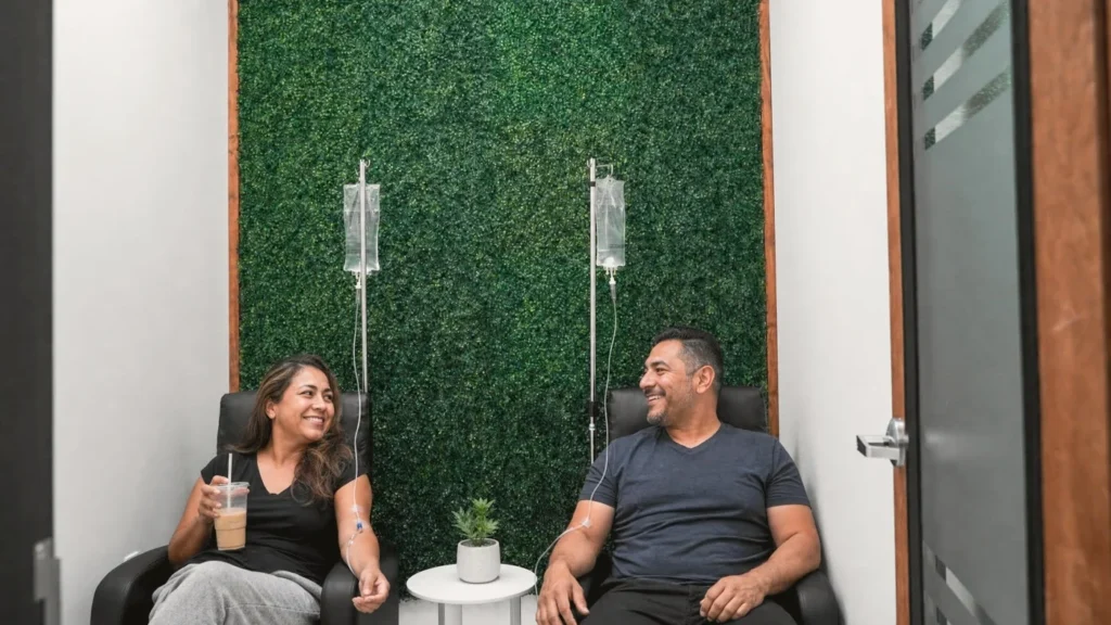 Two adults relax in black recliners with IV drips running, in front of the green plant wall of the IV therapy room at Metropolitan Medical Centers in Tamarac
