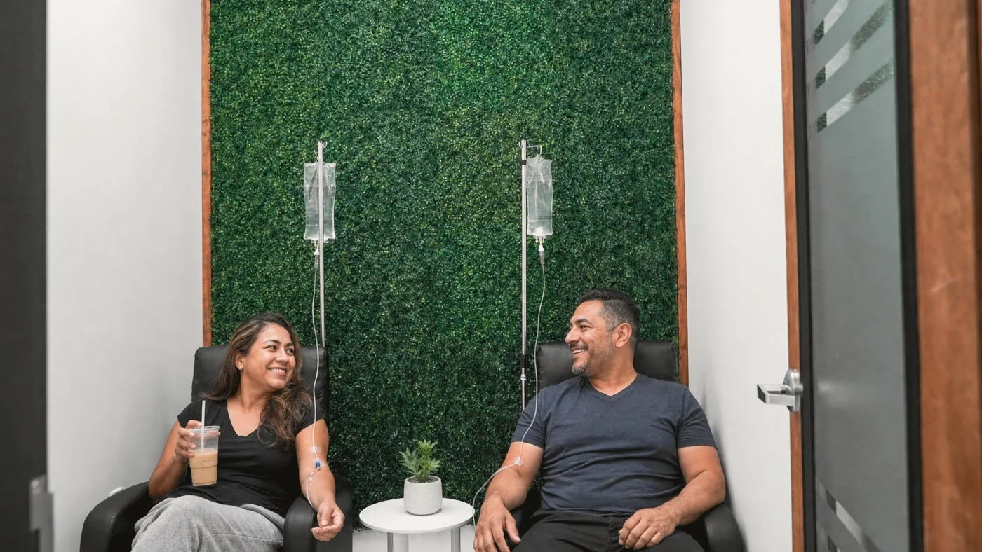 Two adults relax in black recliners with IV drips running, in front of the green plant wall of the IV therapy room at Metropolitan Medical Centers in Tamarac